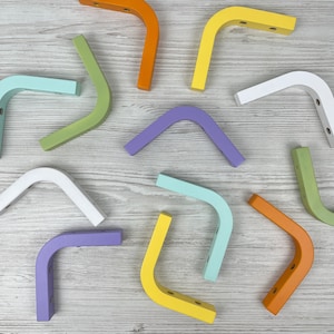 Puede incluir: Una colección de soportes de madera en forma de L, coloridos, en varios tonos pastel, incluyendo naranja, amarillo, verde, azul, morado y blanco. Cada soporte tiene agujeros pre-perforados para el montaje. Los soportes están dispuestos sobre una superficie de madera clara.