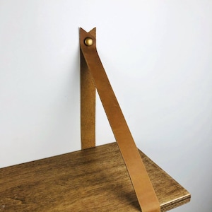 May include: A wooden shelf with a brown leather strap attached to the wall with a brass screw. The shelf is made of natural wood with a light brown stain.