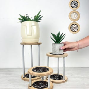 May include: A collection of small plant stands with a modern design. The stands feature a natural wood finish with a black geometric pattern on the top and bottom. The legs are silver. One stand holds a cream-colored face planter with a green plant.