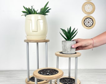 Puede incluir: Un conjunto de pequeños soportes para plantas con un diseño moderno. Los soportes presentan un acabado de madera natural con un patrón geométrico negro. Las patas son plateadas. Un soporte sostiene una maceta con forma de cara crema con una planta verde.
