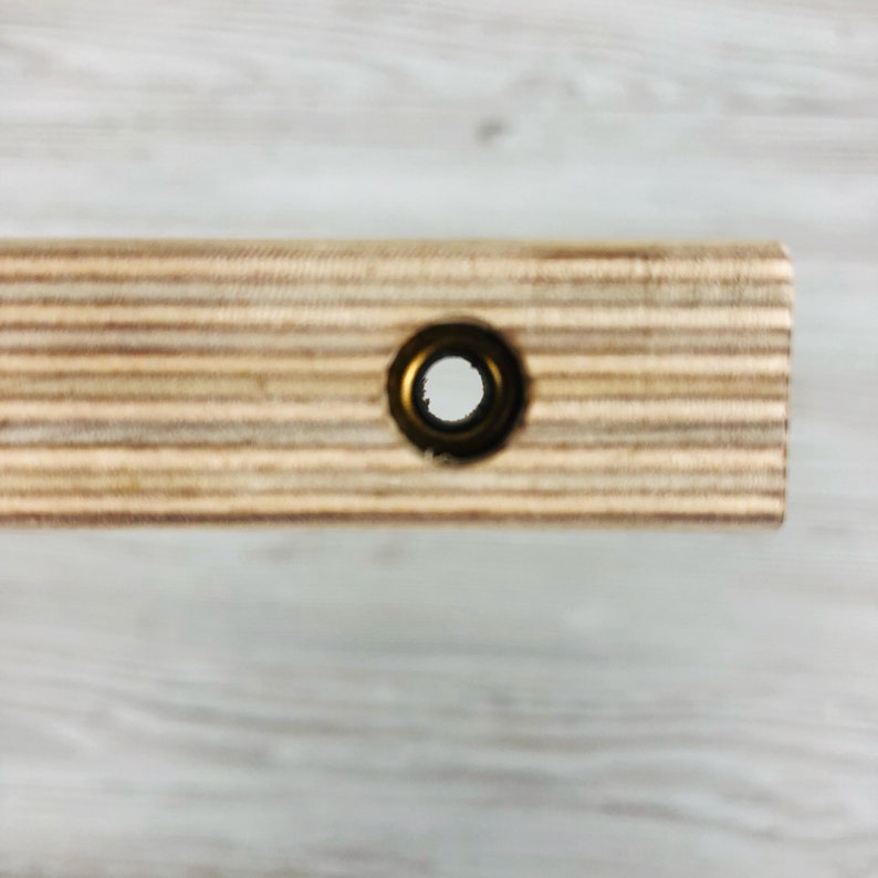Puede incluir: Una pieza rectangular de madera contrachapada de color claro con un patr&oacute;n de veta estratificada visible en el lateral. Un &uacute;nico orificio circular est&aacute; centrado en la superficie plana, reforzado con un ojal met&aacute;lico oscuro.
