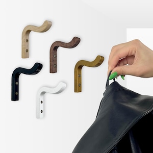 Könnte beinhalten: Eine Sammlung von Wandgarderobenhaken aus Holz in verschiedenen Farben: Naturholz, Dunkelbraun, Gold, Schwarz und Weiß. Die Haken haben ein gebogenes Design. Eine Hand hält eine schwarze Tasche und demonstriert die Verwendung des Hakens.