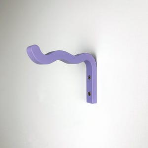 May include: A light purple wall-mounted hook with a wavy design. The hook is made of wood and has two screws.
