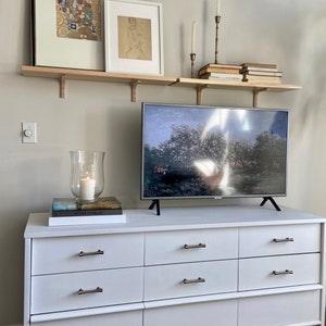 May include: A white dresser with six drawers and brass pulls. The dresser is decorated with a television, two framed pictures, a candle, and books on a shelf above the dresser.