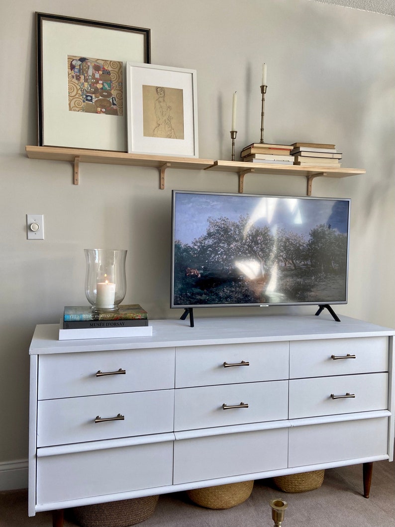 May include: A white dresser with six drawers and brass pulls. The dresser is decorated with a television, two framed pictures, a candle, and books on a shelf above the dresser.