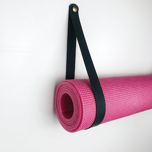 May include: A black leather strap with a brass buckle hangs from a white wall. A pink yoga mat is rolled up and secured to the strap.