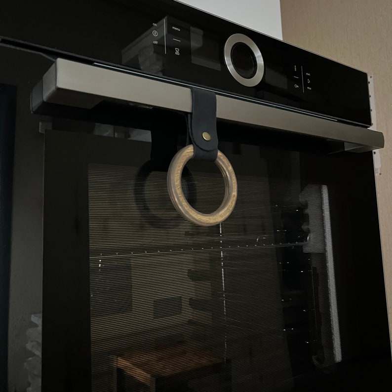 May include: A black oven with a wooden ring attached to the top. The ring is hanging from a black leather strap with a silver buckle.