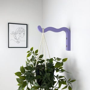 May include: A light purple wooden wall-mounted plant hanger with a curved design. A hanging plant with green leaves is suspended from the hanger.