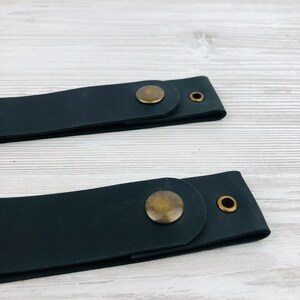 Pu&ograve; includere: Due cinturini in pelle nera con chiusura a bottone in ottone. I cinturini sono su una superficie in legno bianco.