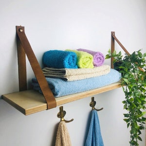 May include: A wooden shelf with brown leather straps and brass hooks. The shelf is holding rolled up towels in shades of blue, green, and purple. A potted plant is hanging off the side of the shelf.
