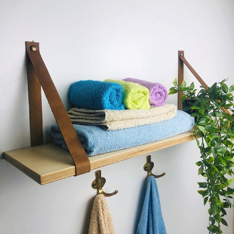 May include: A wooden shelf with brown leather straps and brass hooks. The shelf is holding rolled up towels in shades of blue, green, and purple. A potted plant is hanging off the side of the shelf.