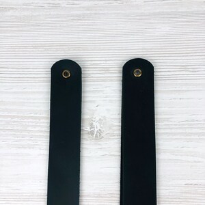 May include: Two black leather straps with brass eyelets. The straps are about 1 inch wide and are shown against a white wood background.