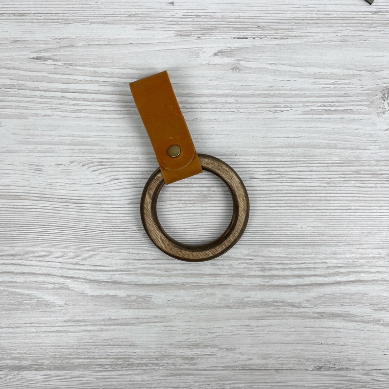 May include: A wooden ring with a brown leather strap. The ring is made of dark wood and has a smooth finish. The strap is attached to the ring with a metal rivet.