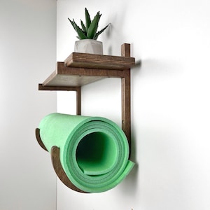 Puede incluir: Una unidad de almacenamiento de madera oscura montada en la pared con un estante superior plano y un soporte inferior curvo. Una pequeña suculenta verde en una maceta geométrica gris descansa en el estante, y una esterilla de espuma verde claro enrollada está en el soporte.