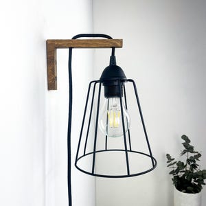 Può includere: Una lampada da parete con una staffa in legno a forma di L color marrone scuro, un paralume a gabbia in metallo nero e un cavo rivestito in tessuto nero. Una lampadina in vetro trasparente con filamenti visibili è sospesa all'interno.