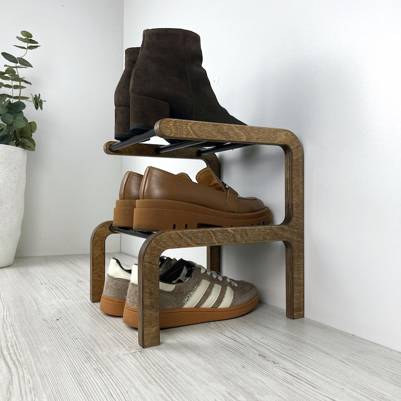 Puede incluir: Un zapatero de madera con tres niveles. El nivel superior tiene un par de botines marrones, el nivel intermedio tiene un par de mocasines marrones y el nivel inferior tiene un par de zapatillas blancas y negras.