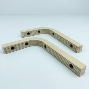 May include: Two wooden shelf brackets with a curved shape and multiple holes for mounting. The brackets are made of light-colored wood with a natural grain.