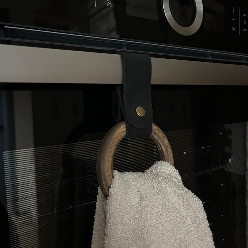 May include: A black leather towel ring with a wooden ring and a brass button. The ring is hanging on a black oven door.