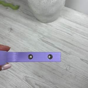 May include: A close-up of a light purple strap with two brass grommets. The strap is made of a smooth, textured material.