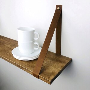 May include: A wooden shelf with a brown leather strap attached to the wall. Two white ceramic mugs are stacked on a white saucer on the shelf.