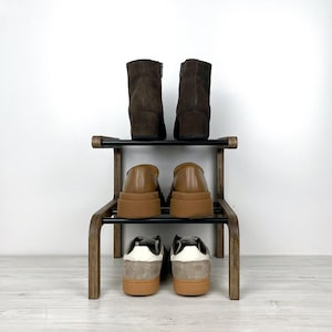 Puede incluir: Un zapatero de madera de dos niveles con estantes de metal negro. El estante superior tiene dos botines marrones y el estante inferior tiene dos pares de zapatillas marrones y grises.