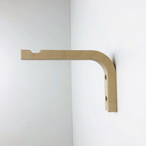Puede incluir: Un soporte de pared de madera de color claro con un dise&ntilde;o en forma de L curva. El brazo horizontal presenta una peque&ntilde;a muesca semicircular en el extremo. La secci&oacute;n vertical tiene dos orificios pretaladrados con tornillos met&aacute;licos visibles.