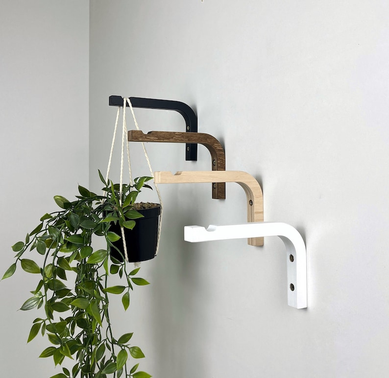 Puede incluir: Cuatro soportes de pared en forma de L montados en fila vertical, con acabados en negro, madera oscura, madera clara y blanco. Cada soporte tiene un extremo con muesca para sujetar un cord&oacute;n. Una maceta negra con una planta verde cuelga del soporte superior.