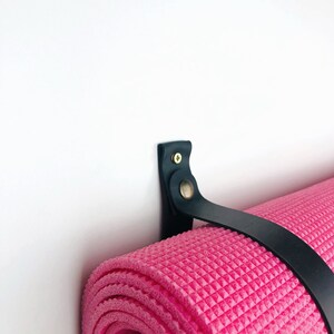 Pu&ograve; includere: Un porta tappetino da yoga in pelle nera montato su una parete bianca. Il porta tappetino ha un cinturino che tiene un tappetino da yoga rosa arrotolato.