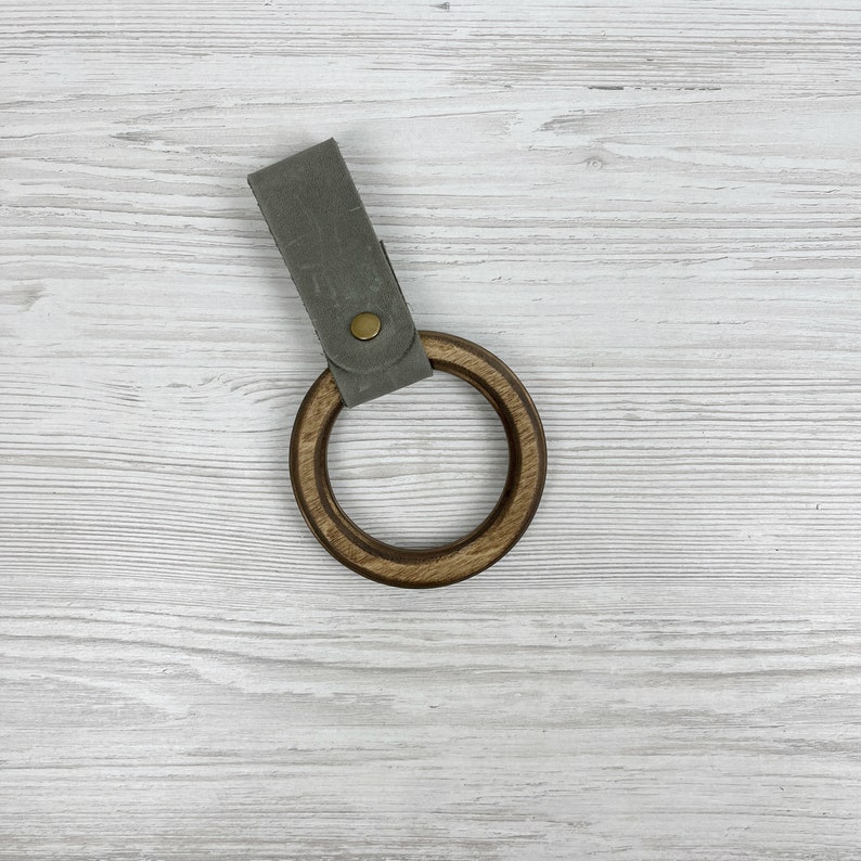 May include: A wooden ring with a gray leather strap. The ring is made of dark wood and has a smooth finish. The strap is attached to the ring with a metal button.
