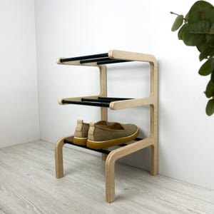 Puede incluir: Un zapatero de tres niveles hecho de madera clara con barras de metal negro. El estante contiene un par de zapatos sin cordones de color beige. El diseño es minimalista y moderno, adecuado para la organización de la entrada o el armario.