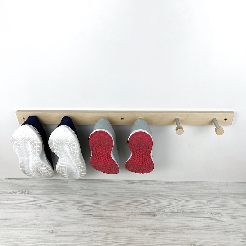 May include: A wooden shoe rack with four hooks for hanging shoes. The rack is mounted on a white wall and has four pairs of shoes hanging from it, including two pairs of white trainers with red accents and two pairs of dark blue trainers.