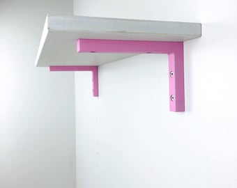 May include: A white shelf with pink brackets mounted on a white wall. The shelf is rectangular and appears to be made of wood. The pink brackets are L-shaped and provide support. The design is modern and minimalist.
