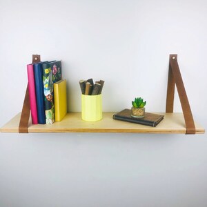 May include: A wooden shelf with brown leather straps, featuring a yellow ceramic container with brown sticks, a small potted succulent, and a stack of books with floral patterns.