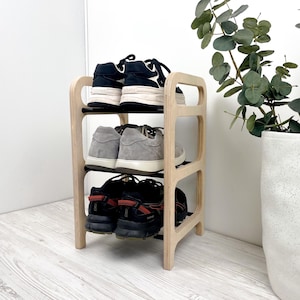 Puede incluir: Un zapatero de madera de tres niveles con un acabado claro. El estante contiene tres pares de zapatos: zapatillas deportivas negras y blancas, zapatillas de ante grises y zapatillas para correr negras con detalles rojos. El estante está junto a una maceta blanca con follaje verde.