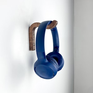 Puede incluir: Un par de auriculares de diadema de color azul oscuro colgados en un gancho de pared de madera marrón oscuro curvado. El gancho está fijado a la pared con dos tornillos metálicos visibles, proporcionando un soporte firme para los auriculares.