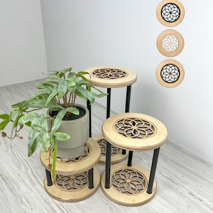Könnte beinhalten: Vier Pflanzenständer aus Holz mit einem lasergeschnittenen Blumendesign auf der Ober- und Unterseite. Die Ständer haben schwarze Metallbeine und sind mit einer Topfpflanze angeordnet. Die Ständer sind aus hellem Holz gefertigt.
