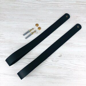 May include: Two black leather straps with gold metal hardware. The straps are laid out on a white wooden surface. There are two gold screws and two silver screws on the surface.