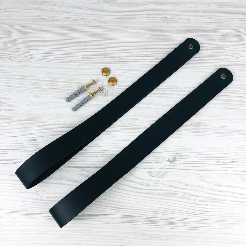 May include: Two black leather straps with gold metal hardware. The straps are laid out on a white wooden surface. There are two gold screws and two silver screws on the surface.