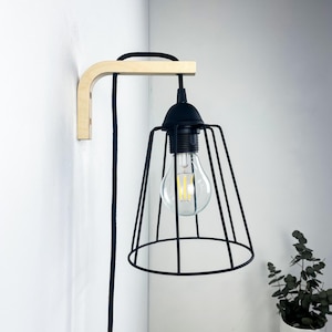 Op de afbeelding: Een moderne wandlamp met een zwarte metalen kooilampenkap en een zichtbare gloeilamp. De lamp wordt ondersteund door een arm van natuurlijk hout en een zwart snoer. Hij is aan een witte muur bevestigd.