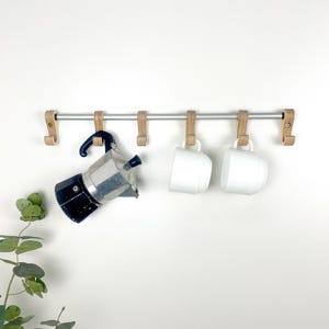 May include: A wall-mounted rack featuring a silver metal rod and six light-colored wooden hooks. Hanging from the hooks are a dark blue and silver metal moka pot, followed by two plain white ceramic mugs, leaving three hooks empty.