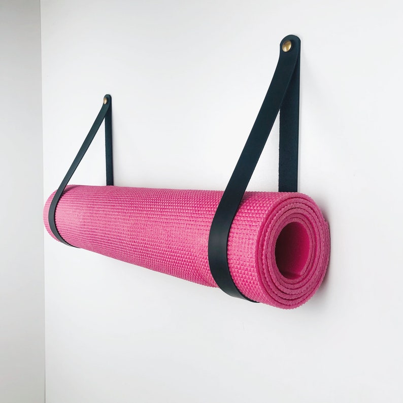 May include: A pink yoga mat hanging on a wall with black leather straps. The straps are attached to the wall with gold screws.