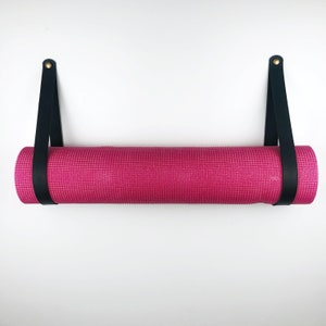 May include: A pink yoga mat hanging on a wall with black leather straps and gold hardware.