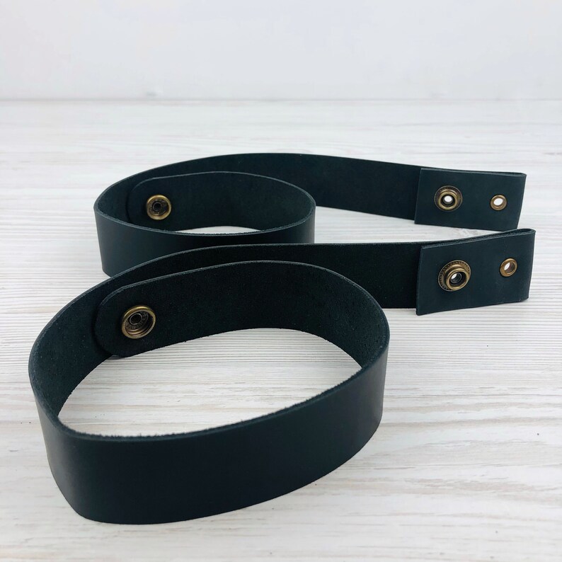 Pu&ograve; includere: Due cinturini in pelle nera con chiusura a scatto in ottone. I cinturini sono larghi circa 2,5 cm e sono mostrati su una superficie di legno bianco.