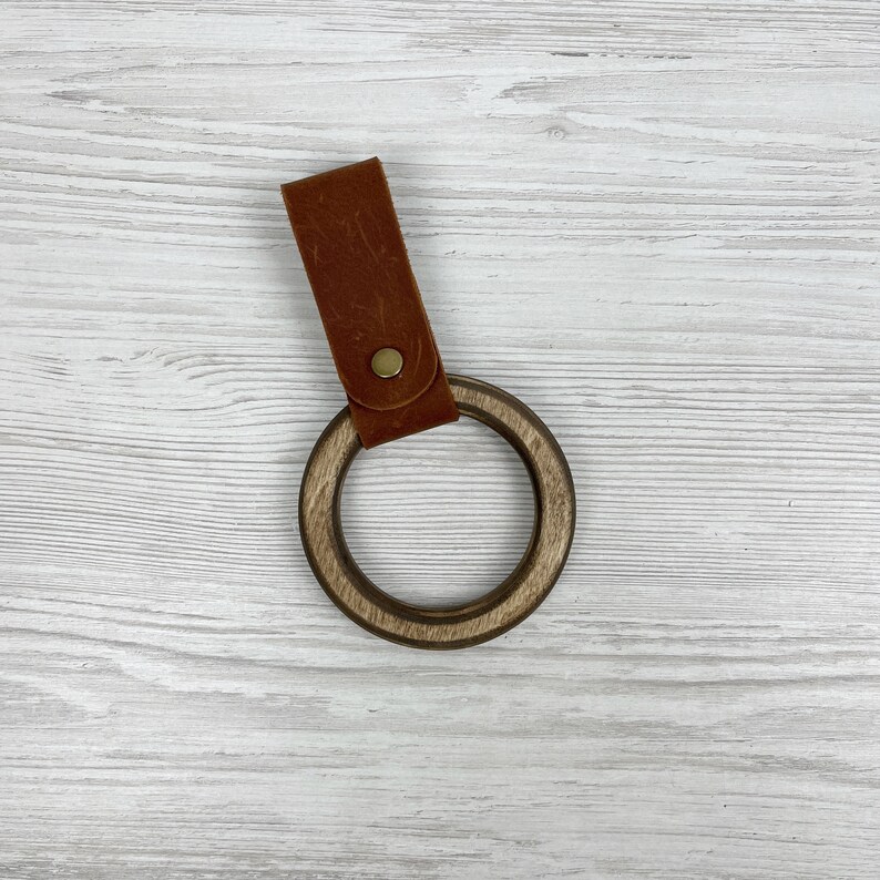 May include: A wooden ring with a brown leather strap. The ring is made of dark wood and has a smooth finish. The strap is attached to the ring with a metal rivet.