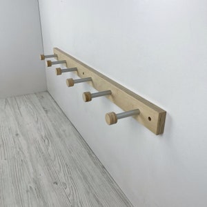 May include: A wooden wall-mounted coat rack with six silver hooks. Each hook has a wooden knob at the end. The rack is made of light-coloured wood and is mounted on a white wall.
