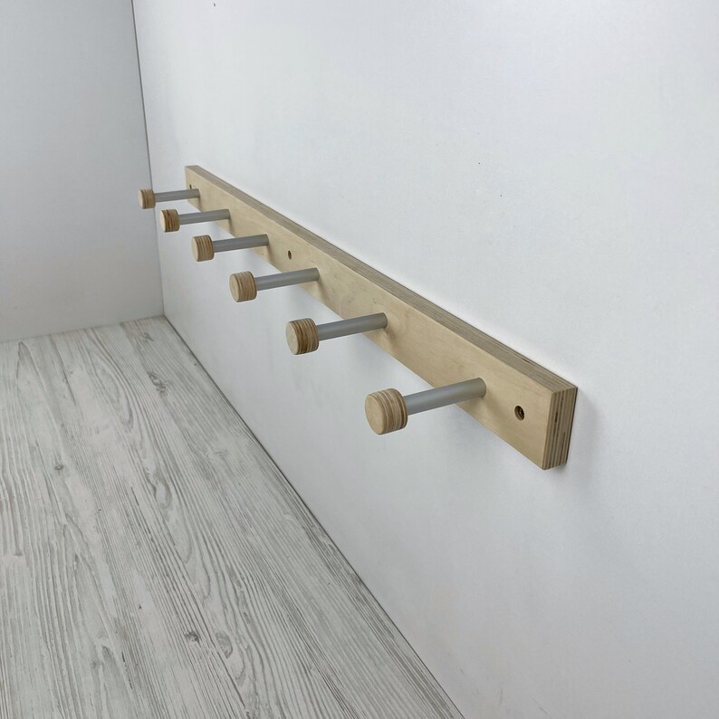 May include: A wooden wall-mounted coat rack with six silver hooks. Each hook has a wooden knob at the end. The rack is made of light-coloured wood and is mounted on a white wall.