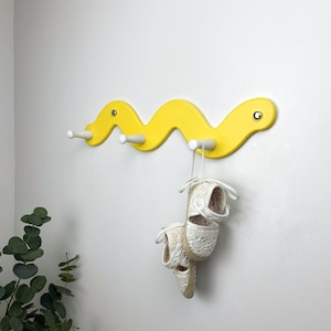 Könnte beinhalten: Eine leuchtend gelbe Wandgarderobe in wellenförmiger Schlangenform mit drei weißen Haken. Ein Paar weiße, gewebte Espadrilles mit Schnürsenkeln hängt am rechten Haken und ist mit den weißen Stoffbändern befestigt.