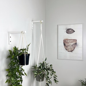 May include: Two white L-shaped metal wall brackets mounted at different heights. Each bracket supports a white macrame plant hanger with a potted green plant. The left plant is in a black pot, and the right plant is in a grey pot.