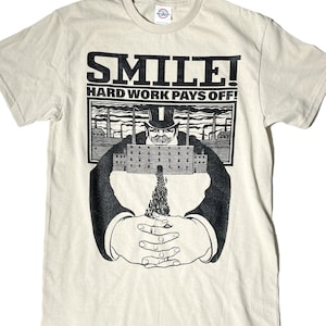 Puede incluir: Una camiseta beige con un estampado gráfico en blanco y negro de un hombre con un sombrero de copa y una fábrica. El texto "SMILE! HARD WORK PAYS OFF!" está impreso sobre el gráfico.