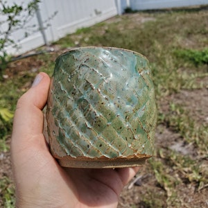 May include: A handmade ceramic tumbler with a textured, diamond-patterned exterior. The cup features a blend of green and turquoise glazes, with a natural, unglazed base. The design is rustic and organic, ideal for enjoying beverages.